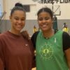 Two women posing together on a basketball court, one wearing a green jersey and the other in a brown sweatshirt.