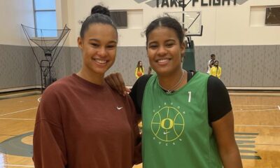 Two women posing together on a basketball court, one wearing a green jersey and the other in a brown sweatshirt.