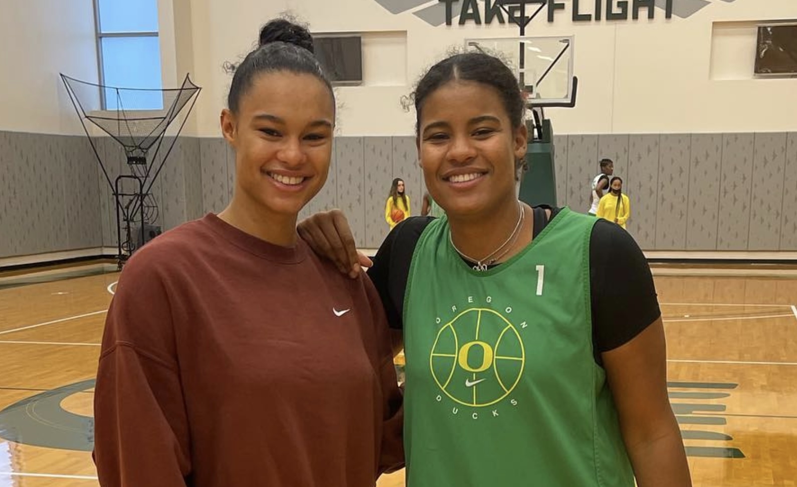 Two women posing together on a basketball court, one wearing a green jersey and the other in a brown sweatshirt.